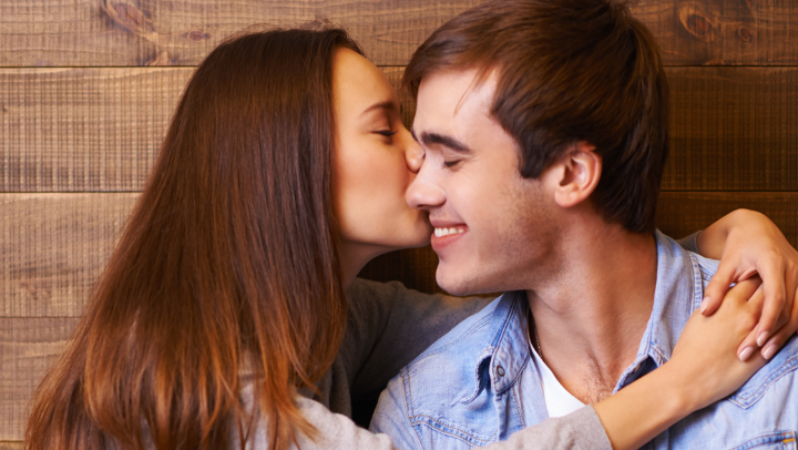 Zwei Junge Erwachsene, eine Frau und ein Mann, sitzen nebeneinander. Die Frau umarmt den Mann und gibt ihm einen Kuss auf die Wange.