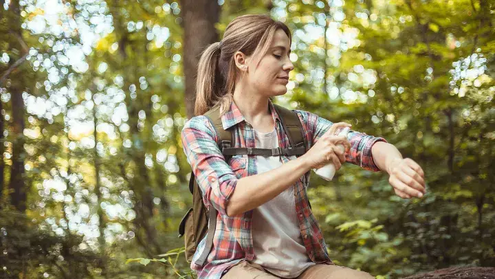 Junge Frau im Wald mit einem Wanderrucksack auf dem Rücken, besprüht gerade ihren linken Arm mit einer Flüssigkeit.