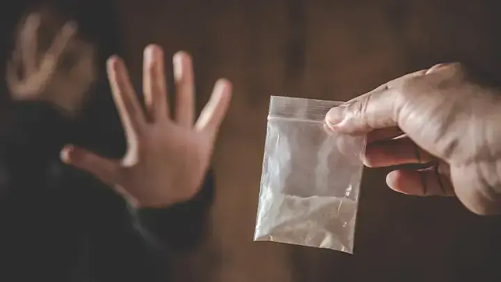 Im Vordergrund eine Hand, die ein Tütchen mit weißem Pulver hält. Im Hintergrund ist schemenhaft eine Person zu sehen, die abwehrend ihre linke Hand nach vorne gegen das Tütchen streckt.