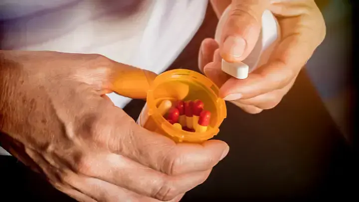 Zwei Hände, die eine Dose mit rot-gelben kapselförmigen Tabletten halten.