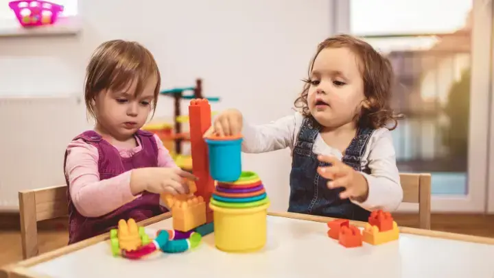Zwei Kleinkinder sitzen an einem Kindertisch und spielen mit Stapelbechern und Bausteinen.