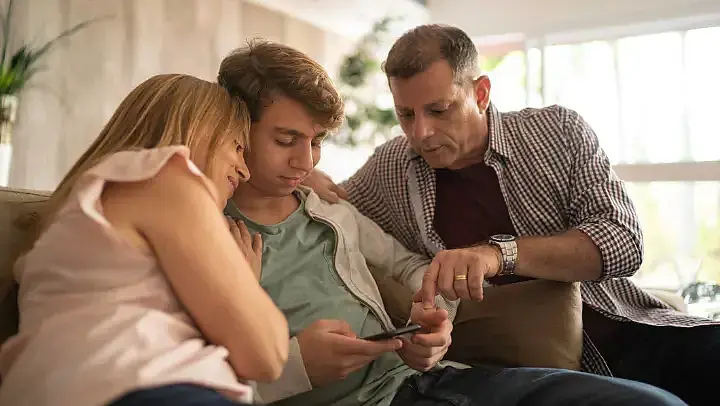 Eltern sitzen mit ihrem jugendlichen Sohn auf dem Sofa und schauen gemeinsam auf ein Handy, das der Sohn in der Hand hält.