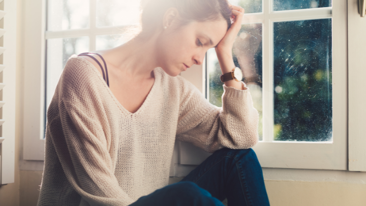 Junge Frau sind vor einem Fenster. Sie stützt den Kopf auf eine Hand, während sie traurig nach unten schaut.