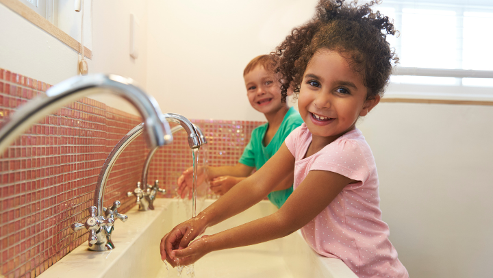Zwei Kinder stehen an einem großen Waschbecken mit drei Wasserhähnen und halten ihre Hände unter fließendes Wasser. Beide lächeln.