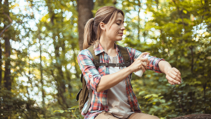 Junge Frau im Wald mit einem Wanderrucksack auf dem Rücken, besprüht gerade ihren linken Arm mit einer Flüssigkeit.