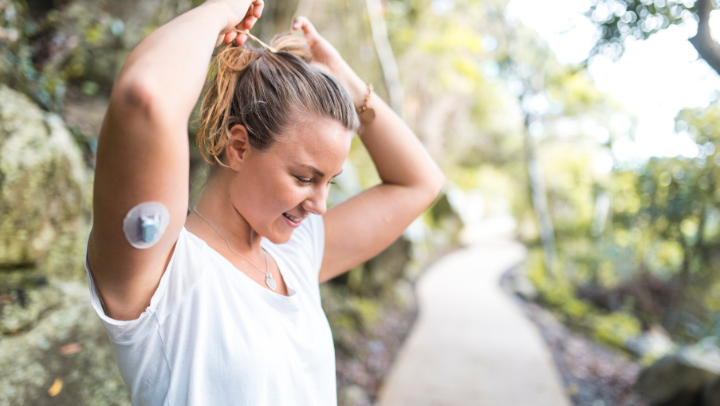 Junge Frau steht auf einem Waldweg und bindet sich die Haare hoch. Am Oberarm sieht man einen Glukosesensor.