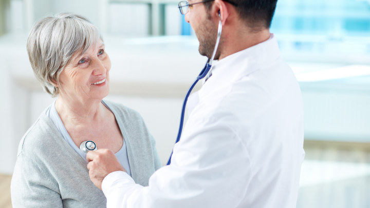 Ältere Frau wird von einem Arzt mit dem Stethoskop untersucht. Sie blickt ihn lächelnd an.